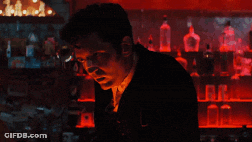 A Man In A Suit Stands In Front Of A Bar With Bottles Of Alcohol On Shelves GIF