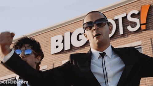 A Man In A Suit Stands In Front Of A Big Ots Sign GIF