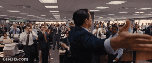 A Man In A Suit Stands In Front Of A Crowd Of People In An Office With A Sign On The Wall That Says " Stocks " GIF