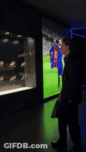 A Man In A Suit Stands In Front Of A Display Case With Soccer Trophies And A Shirt That Says Messi GIF