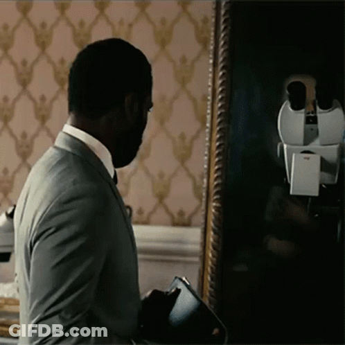 A Man In A Suit Stands In Front Of A Mirror Looking At His Reflection GIF