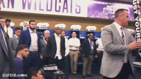 A Man In A Suit Stands In Front Of A Sign That Says " Wildcats " GIF