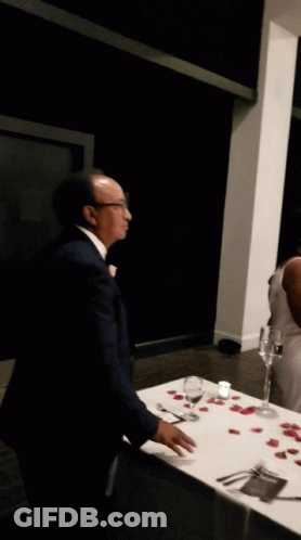 A Man In A Suit Stands In Front Of A Table With Wine Glasses And Rose Petals On It GIF