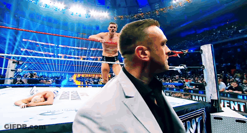 A Man In A Suit Stands In Front Of A Wrestling Ring That Says Saturday Nights Mat GIF