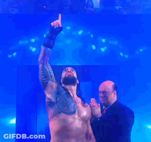 A Man In A Suit Stands Next To A Shirtless Wrestler Who Is Pointing Up At The Sky GIF