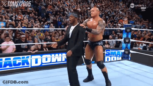 A Man In A Suit Stands Next To A Wrestler In A Wrestling Ring That Says Back Down GIF