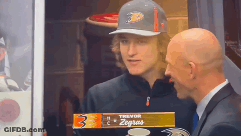 A Man In A Suit Talks To A Young Man Wearing A Hat That Says Trevor Zegras GIF