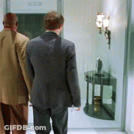 A Man In A Suit Walks With Another Man In A Tan Jacket GIF