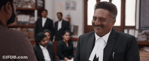A Man In A Suit With The Letter M On His Lapel Smiles GIF