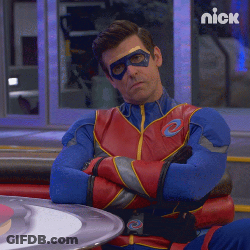 A Man In A Superhero Costume Is Sitting At A Table With His Arms Crossed And A Nick Logo Behind Him GIF