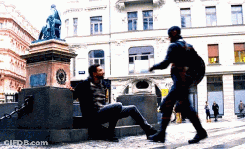 A Man In A Superhero Costume Is Talking To A Man Sitting On The Ground GIF