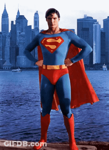 A Man In A Superman Costume Stands In Front Of A City Skyline With His Hands On His Hips GIF