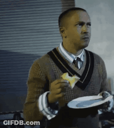 A Man In A Sweater And Tie Is Holding A Plate Of Food And Eating A Piece Of Cheese . GIF