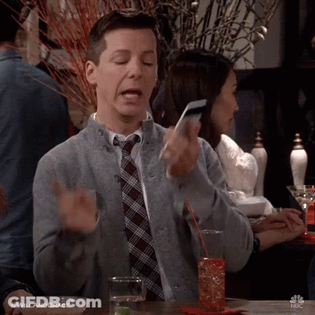 A Man In A Sweater And Tie Is Sitting At A Table Holding A Cell Phone . GIF