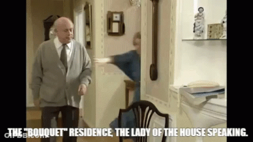 A Man In A Sweater And Tie Is Standing In A Living Room Next To A Woman In A Blue Robe . GIF