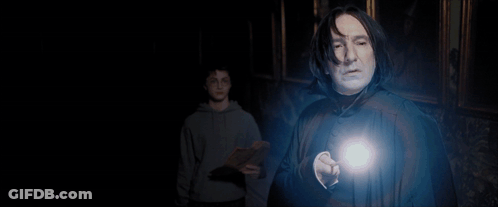 A Man In A Sweater And Tie Stands In A Dark Room With A Light Shining On Him GIF