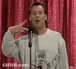 A Man In A Sweater Is Giving A Speech In Front Of A Microphone And Pointing At The Camera . GIF