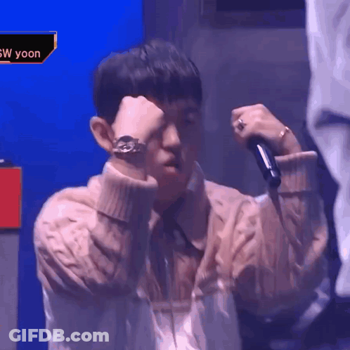 A Man In A Sweater Is Holding A Microphone And Covering His Face . GIF
