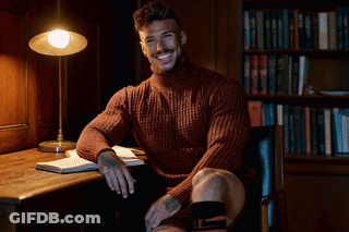 A Man In A Sweater Is Sitting At A Desk With A Lamp GIF