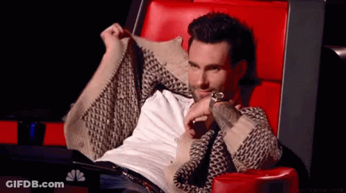 A Man In A Sweater Is Sitting In A Red Chair And Holding His Sweater Over His Shoulder . GIF