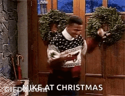 A Man In A Sweater Is Standing In Front Of A Door Holding A Wreath And Says `` Mike At Christmas '' GIF