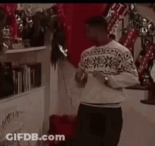 A Man In A Sweater Is Standing In Front Of A Fireplace In A Room . GIF