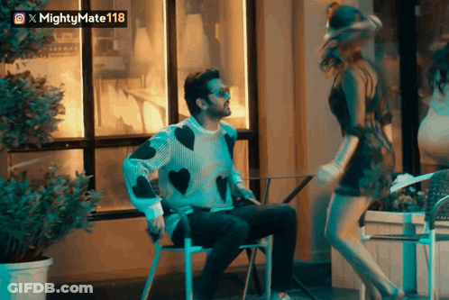 A Man In A Sweater With Hearts On It Sits Next To A Woman In A Short Dress GIF