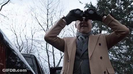 A Man In A Tan Coat Looks Through Binoculars In Front Of A Car GIF