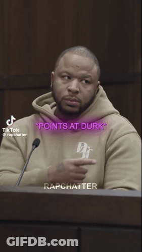 A Man In A Tan Hoodie Points At The Words Points At Durk On His Shirt GIF