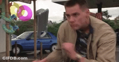 A Man In A Tan Jacket And Tie Is Sitting In Front Of A Blue Car And Inflatable Donuts . GIF