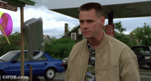 A Man In A Tan Jacket Stands In Front Of A Gas Station With A Sign That Says 113 GIF