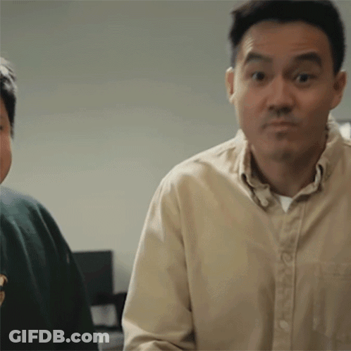 A Man In A Tan Shirt Is Making A Funny Face While Standing Next To Another Man In A Green Sweater . GIF
