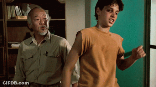 A Man In A Tan Shirt Stands Next To Another Man In A Yellow Shirt GIF