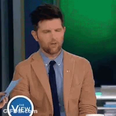 A Man In A Tan Suit And Tie Is Holding A Piece Of Paper In Front Of A Sign That Says The View . GIF