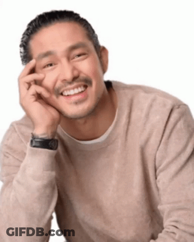 A Man In A Tan Sweater Is Smiling With His Hand On His Chin GIF