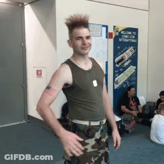 A Man In A Tank Top And Camo Pants Is Standing In Front Of A Welcome To Comic Con International Sign GIF