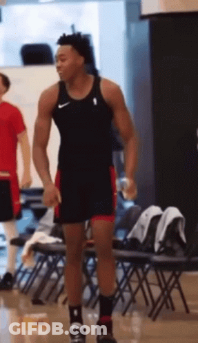 A Man In A Tank Top And Shorts Is Standing In Front Of A Row Of Folding Chairs . GIF