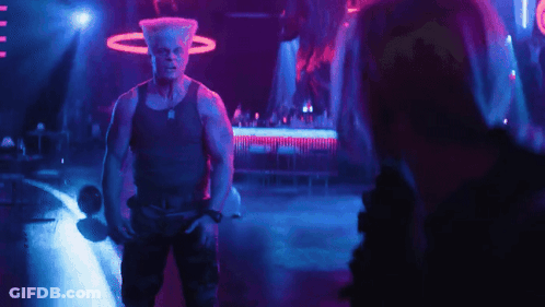 A Man In A Tank Top Stands In A Dark Room GIF