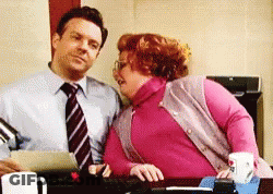 A Man In A Tie And A Woman In A Pink Sweater Are Sitting At A Desk Looking At Each Other GIF