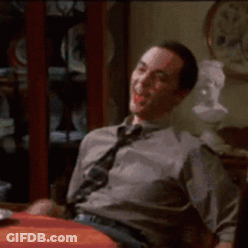 A Man In A Tie Is Sitting At A Table With A Clock In The Background GIF