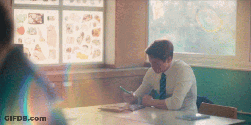 A Man In A Tie Sits At A Desk In A Classroom Looking At His Phone GIF
