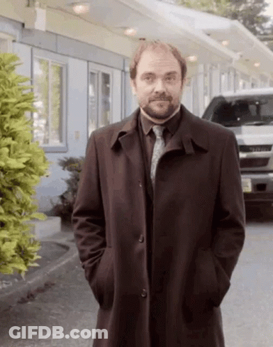 A Man In A Trench Coat And Tie Is Standing In Front Of A Motel . GIF