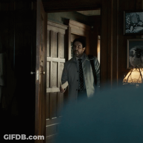 A Man In A Trench Coat And Tie Stands In A Hallway GIF