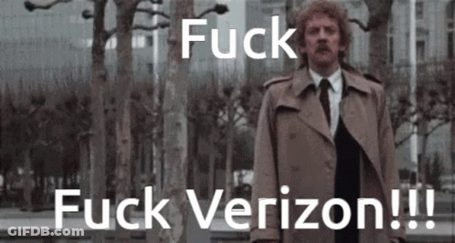 A Man In A Trench Coat And Tie Stands In Front Of Trees And Says Fuck Verizon !!! GIF