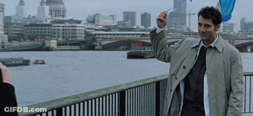 A Man In A Trench Coat And Tie Stands On A Bridge Overlooking A River GIF