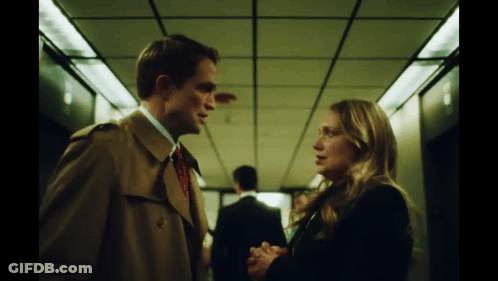 A Man In A Trench Coat And Tie Talks To A Woman GIF