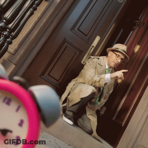 A Man In A Trench Coat Is Kneeling Down In Front Of A Door And Pointing At The Time GIF