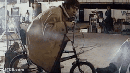 A Man In A Trench Coat Is Riding A Bicycle With The Number 64 On The Front Wheel GIF
