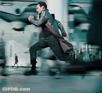 A Man In A Trench Coat Is Running Down A City Street GIF