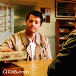 A Man In A Trench Coat Is Sitting At A Table Talking To Another Man In A Room GIF